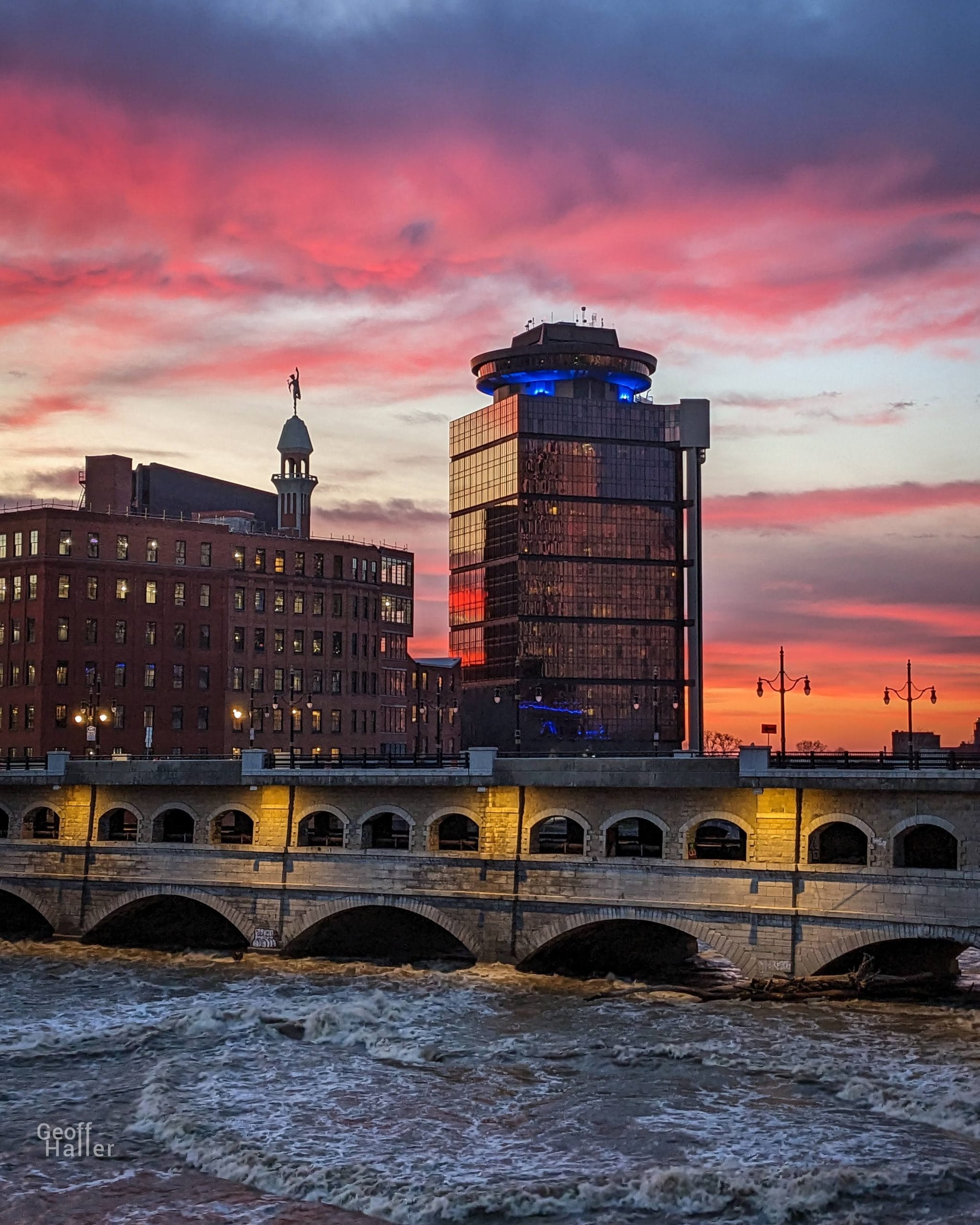 A Rochester cityscape at sunset with vibrant pink and orange clouds, a tall modern glass building, an older brick building with a statue, and a lit up stone bridge spanning the choppy river below.