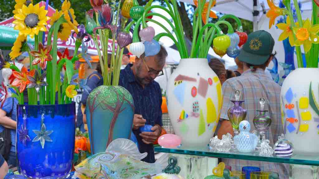 A vibrant display of colorful glass art, featuring vases and flowers, is showcased at Crafts at Lyndhurst. A man in a blue shirt examines items while others browse the collection. Trees and vendor tents are visible in the background, setting a picturesque scene.