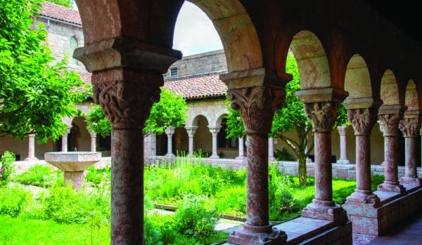 A view through stone arches of a medieval-style cloister garden at The Met Cloisters in Inwood with lush greenery, stone columns, and a central fountain, nestled among old stone buildings and tiled roofs in New York City.