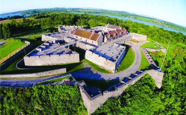 Aerial view of a large stone fort surrounded by lush green trees, with a river visible in the background; the fort has thick walls and a courtyard in the center.