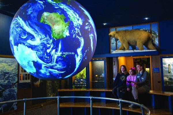 Three people sit on a bench in a museum exhibit with a large illuminated globe of Earth and a stuffed polar bear in the background. The dimly lit room highlights natural history displays, evoking the geological wonders of the Finger Lakes region.