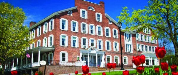 The The Middlebury Inn, a historic red-brick building with white shutters and trim, surrounded by blooming red tulips and green trees under a bright blue sky.