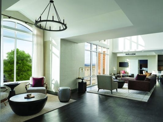 Modern Hudson Valley lodging at Wm Farmer and Sons in Hudson New York with large windows, neutral furniture, dark wood floors, and abundant natural light. Features armchairs, a round coffee table, a sofa, and scenic views of trees and mountains outside.