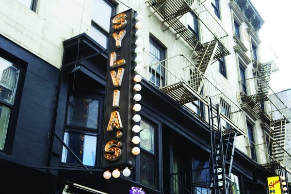A vintage-lit sign reading SYLVIAS hangs vertically on the exterior of a city building with fire escapes, evoking the classic charm of a New York City restaurant in an urban street setting.