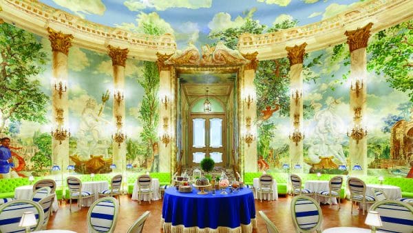 Lavish New York City dining room with tall marble columns, ornate chandeliers, and colorful murals of gardens and classical scenes. Round tables with blue and white chairs and a central blue-draped table are set for tea.