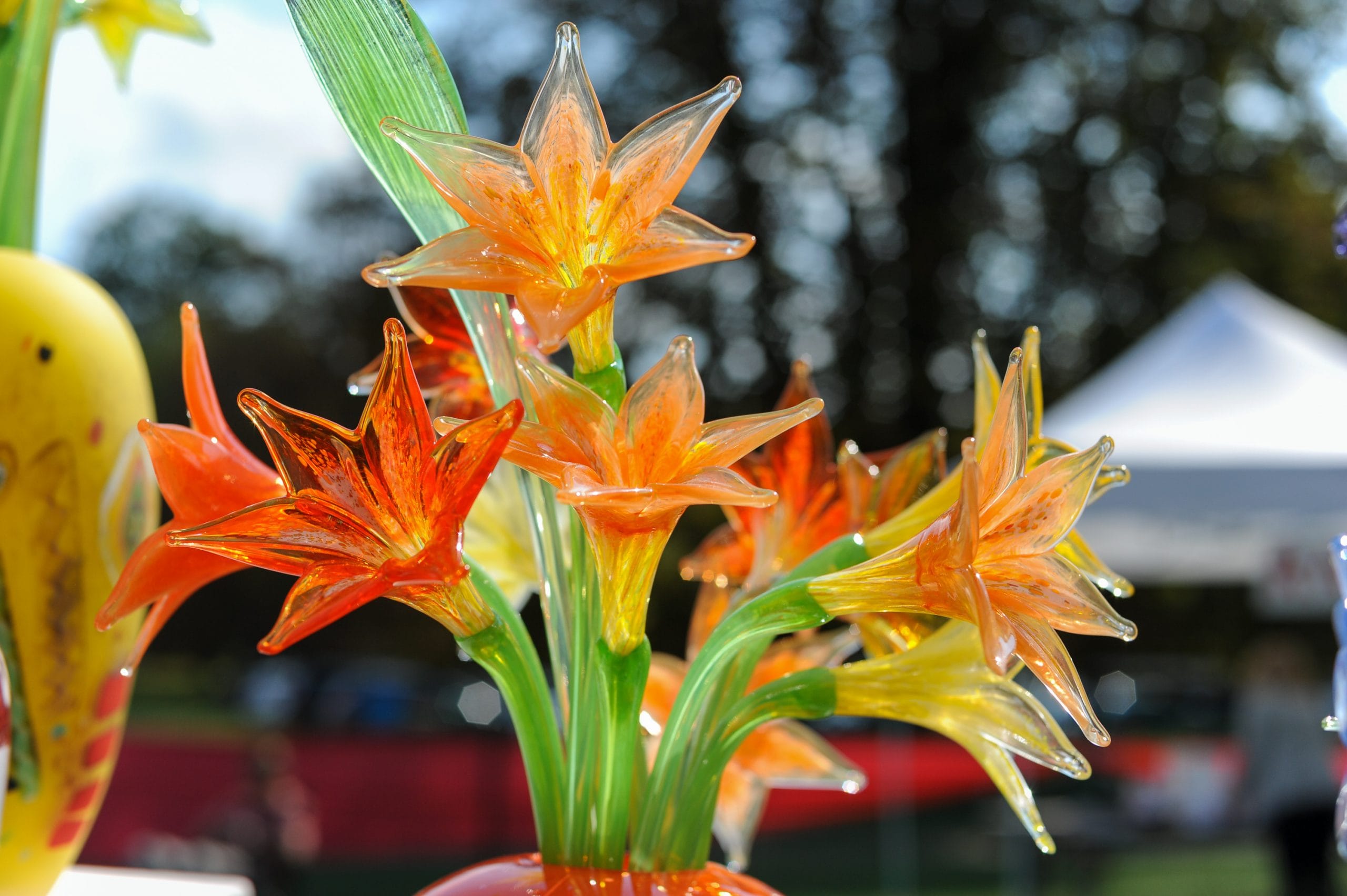 Close-up of vibrant glass flowers in shades of orange and yellow, intricately crafted with green stems and leaves, reminiscent of the stunning crafts at Lyndhurst. The blurred background shows trees and a white tent under a blue sky.