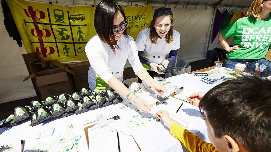 People at a booth wearing white Car Free NYC shirts interact with a visitor, celebrating Earth Day. The table is covered with papers and promotional items. A yellow banner with transportation symbols is in the background, emphasizing sustainable living.