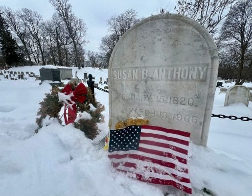 A snowy cemetery in Rochester with Susan B. Anthony’s gravestone in the foreground, adorned with a wreath and an American flag. Other tombstones and bare trees stand quietly in the background.