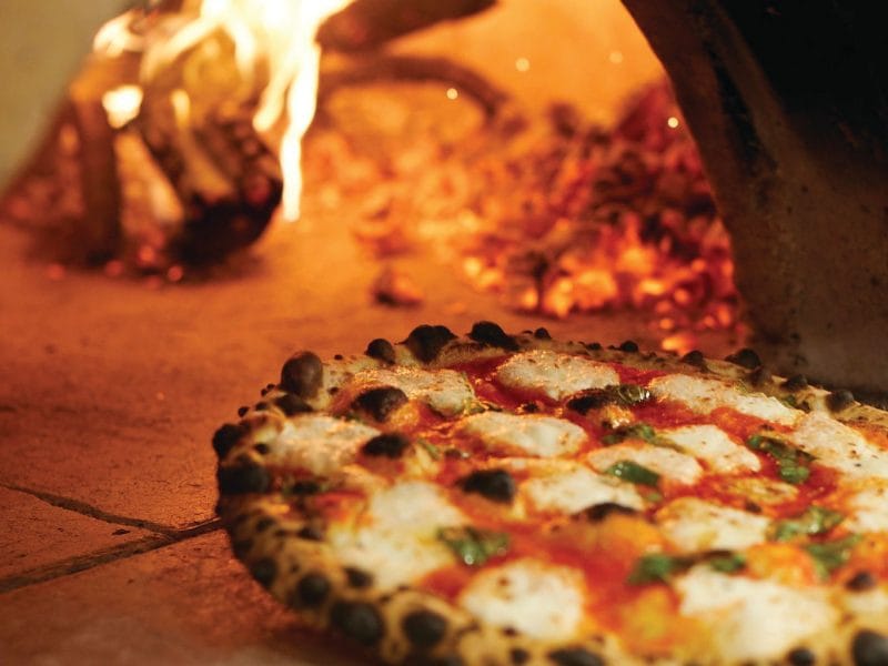 A wood-fired pizza with melted cheese, tomato sauce, and basil leaves sits on a peel in front of a blazing fire inside a traditional brick oven—an iconic stop on any New York food tour for true Empire State Eats.