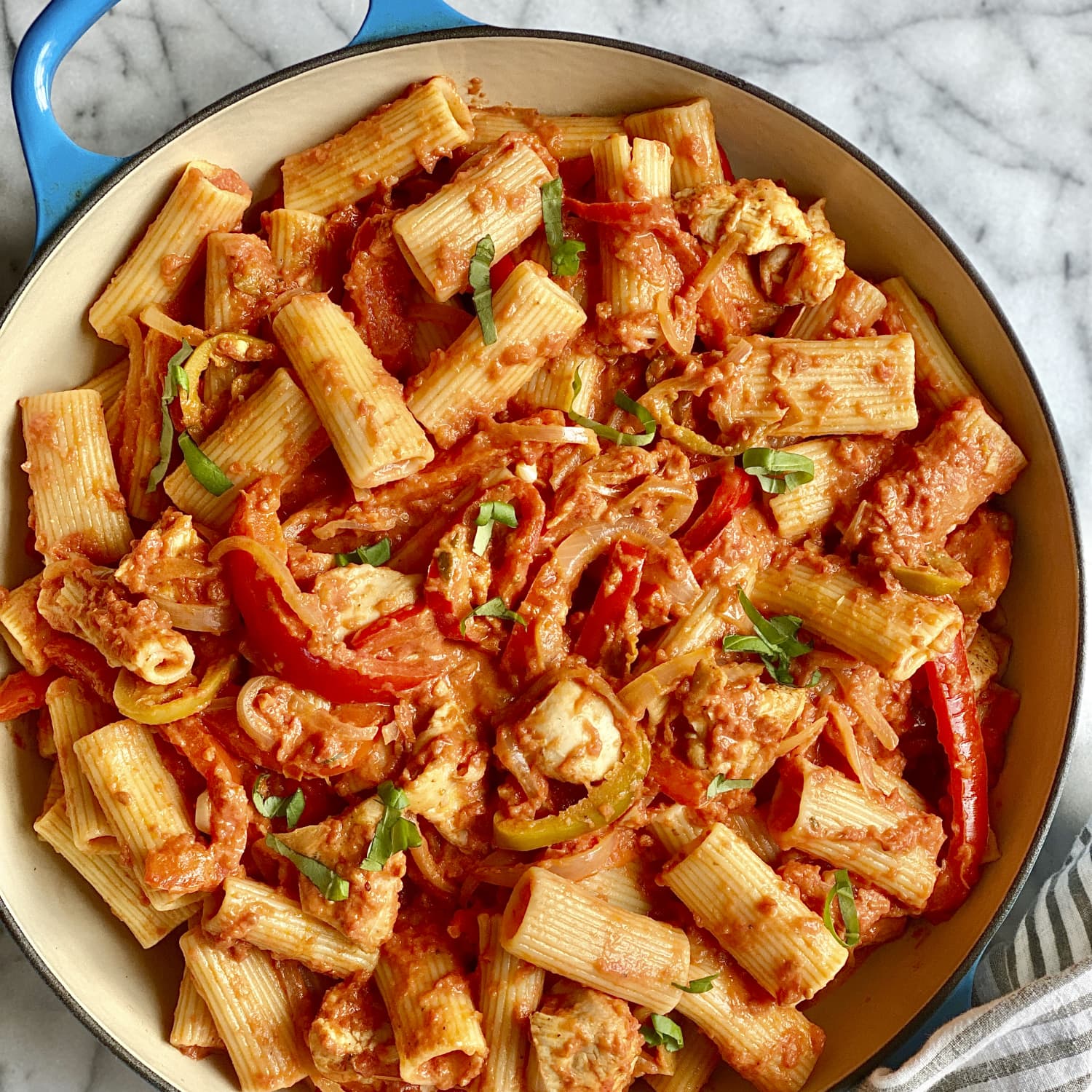 A skillet filled with rigatoni, sliced bell peppers, and chunks of chicken in creamy tomato sauce, garnished with basil—a taste reminiscent of Empire State eats on a marble countertop.