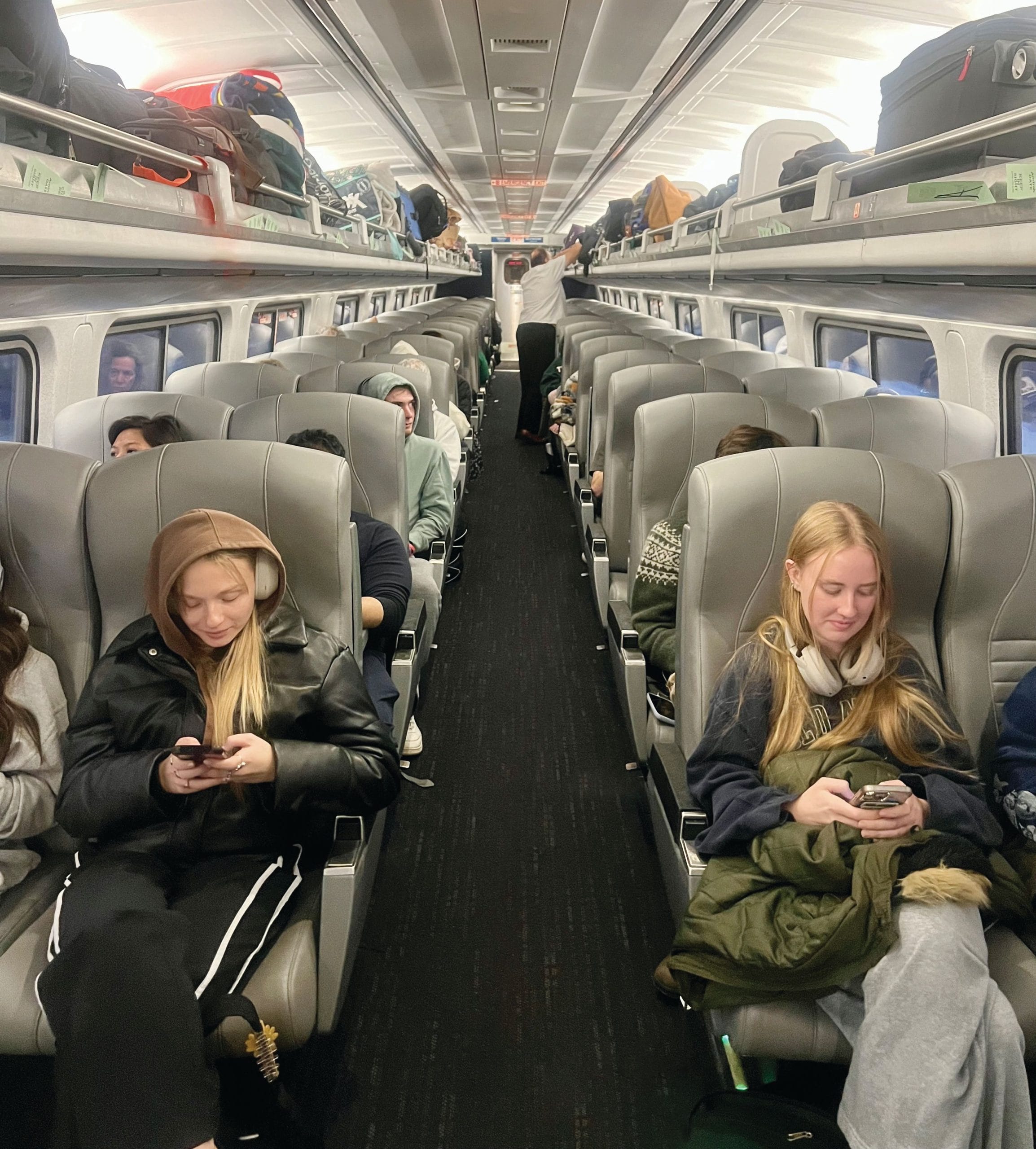 Passengers sit in gray seats inside a modern Rochester train, with luggage stored overhead. Most people are looking at their phones, and the aisle runs down the center of the brightly lit carriage.