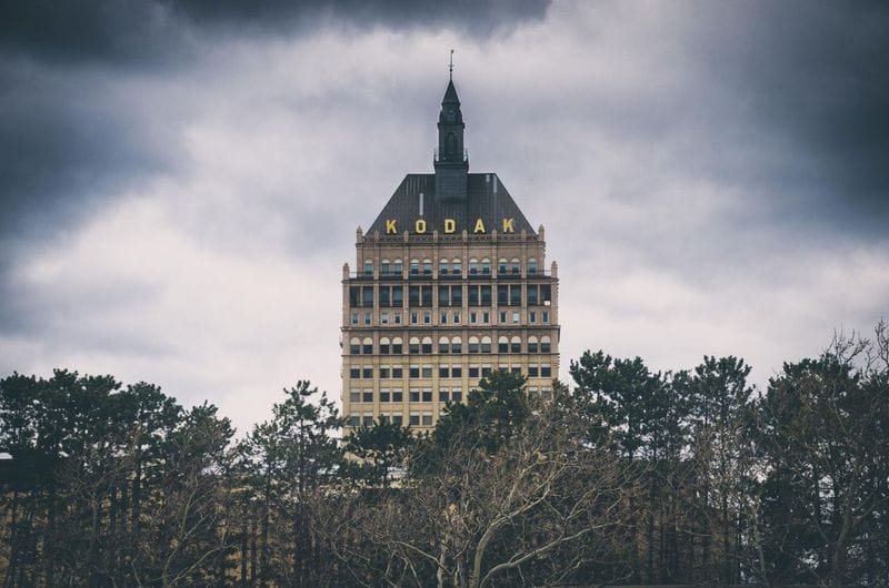 A tall, historic building with KODAK in yellow letters near the top stands behind leafless trees under a cloudy, overcast Rochester sky.