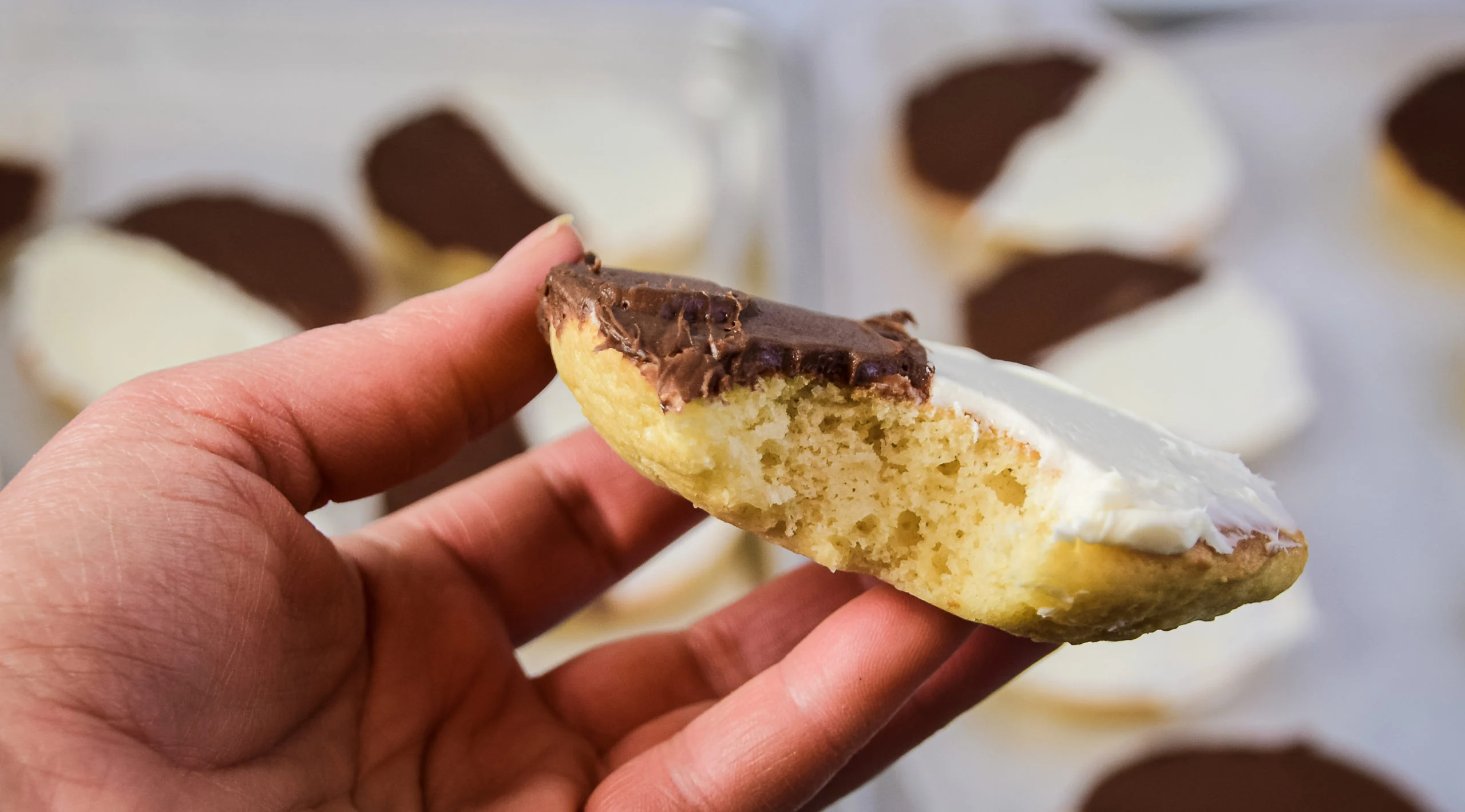 A hand holds a half-eaten black and white cookie, an iconic treat among New York regional dishes, topped with chocolate icing on one side and white icing on the other. More cookies are blurred in the background.