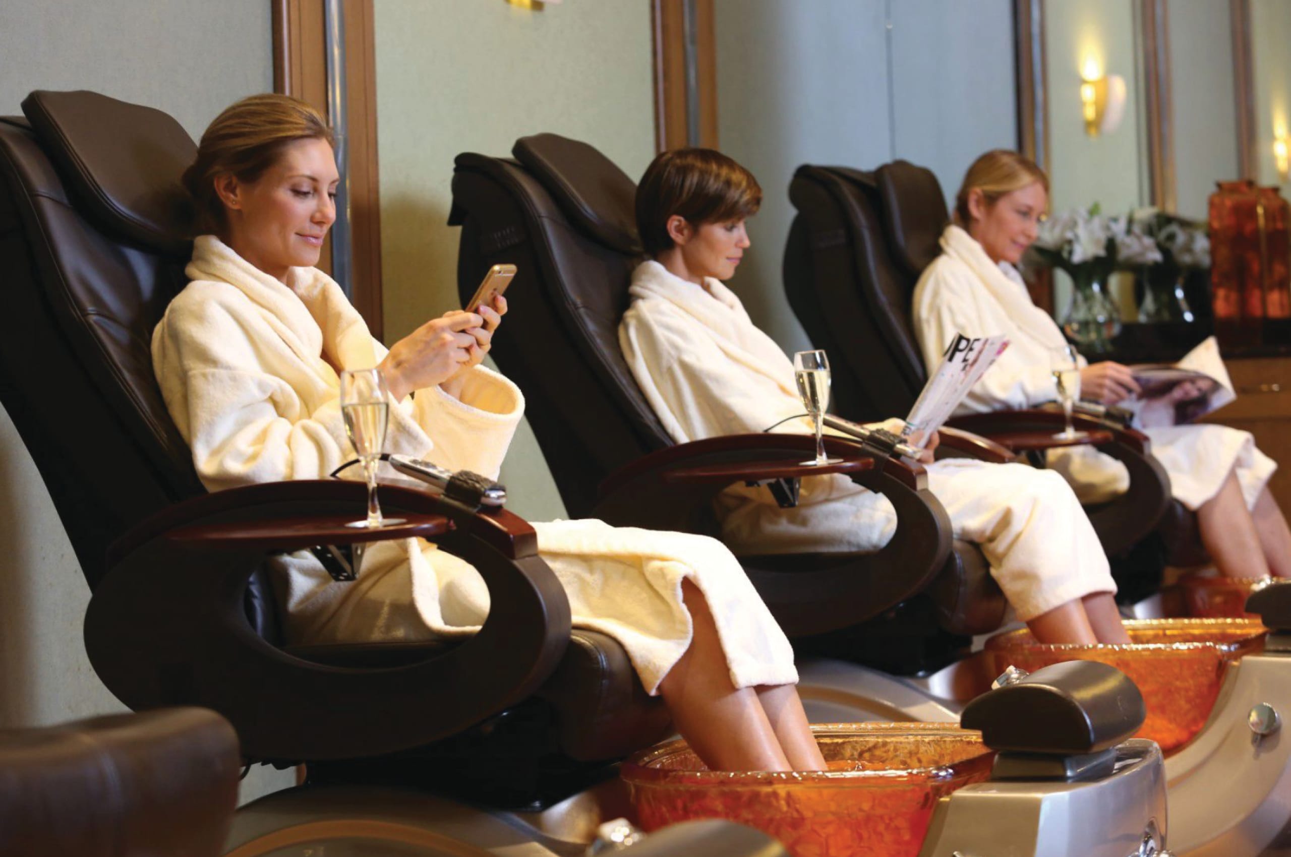 Three women in white robes enjoy relaxation at a luxury spa, sitting in massage chairs with their feet soaking in foot baths. One uses her phone, another reads a magazine, while the third gazes ahead. A glass of champagne rests on a side table.