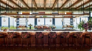 A stylish bar at Klocke Estate features floral upholstered stools, a wooden counter, shelves of colorful bottles, hanging pendant lights, and large windows with scenic views. A bouquet of purple flowers sits at one end of the bar.