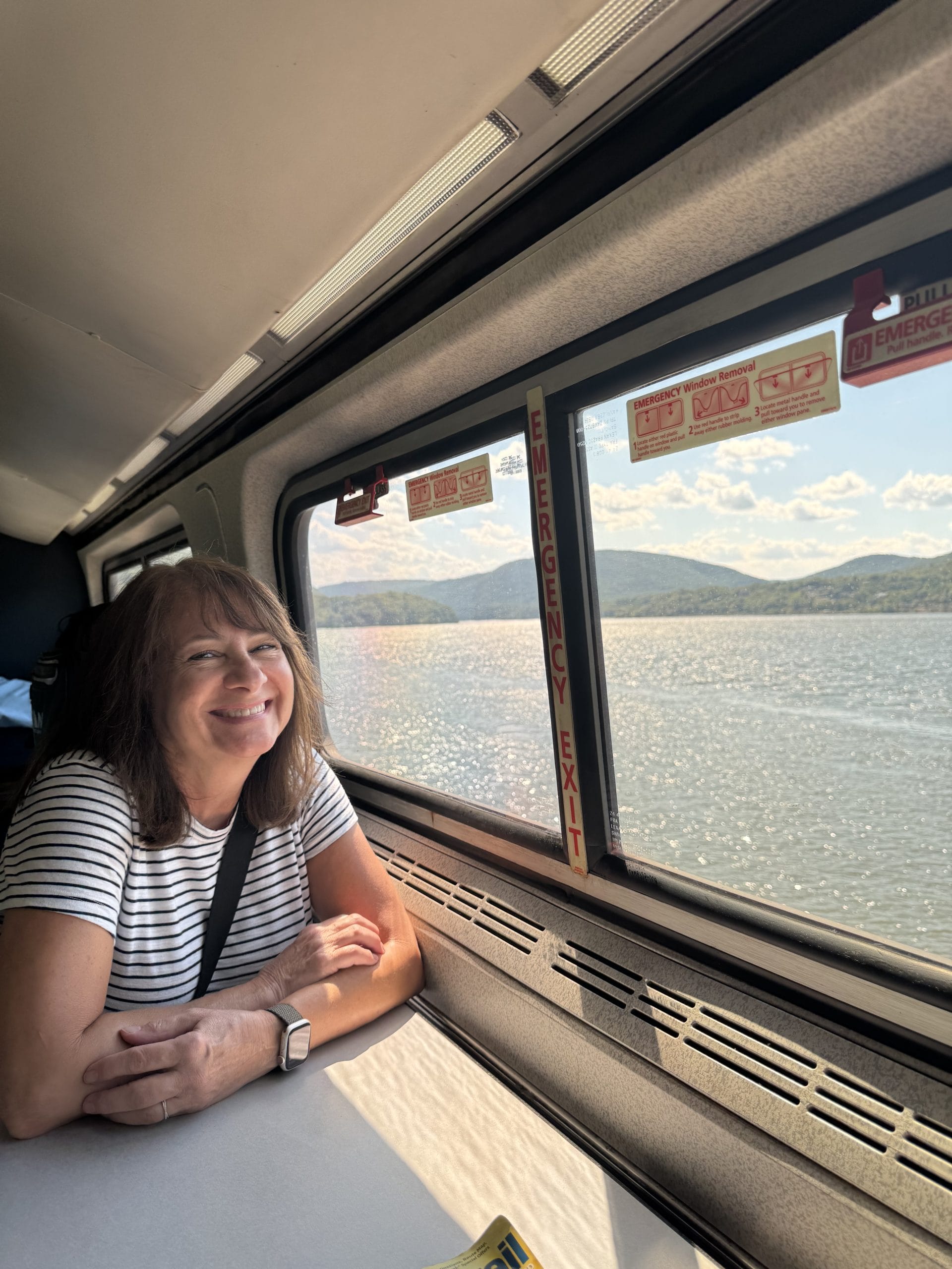 A woman with shoulder-length brown hair and a striped shirt smiles while sitting at a table on a train, ready to explore Upstate New York. Out the window, a calm lake and green hills appear under a partly cloudy sky.
