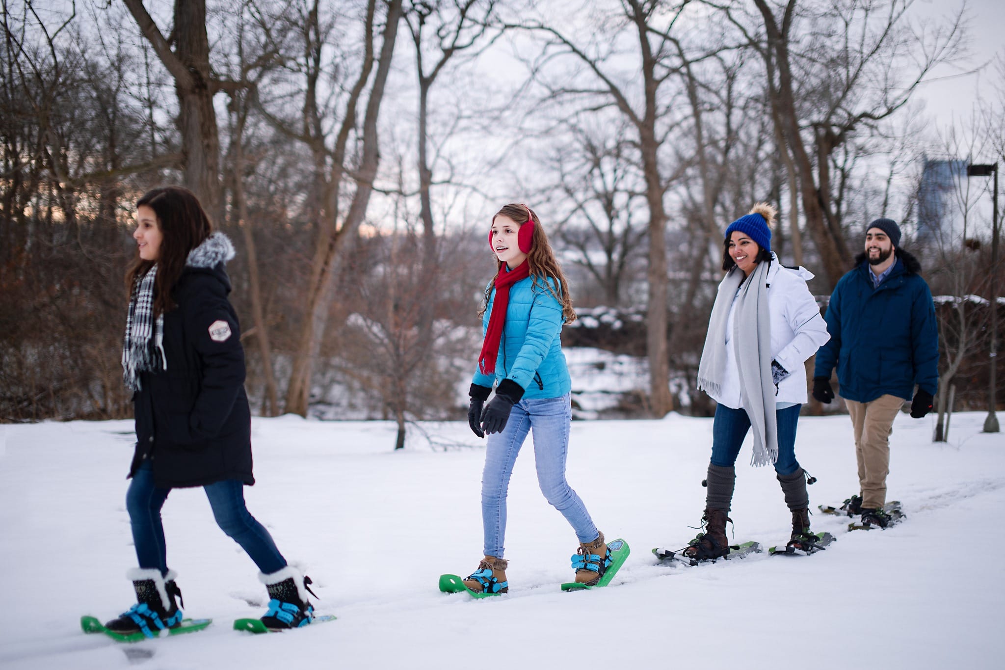 Four people walk in a line through a snowy, wooded area at Jingle Falls USA, wearing snowshoes and winter clothing, smiling and enjoying the winter outdoors.
