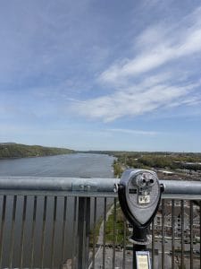 A coin-operated binocular viewer is mounted on a railing overlooking a wide river—an inviting stop on a Walking Tour of Poughkeepsie, with green hills to the left, city buildings to the right, and a blue sky with scattered clouds above.