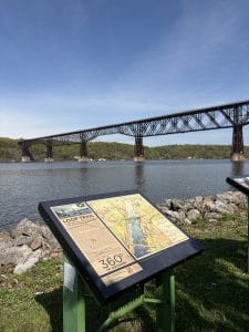 An informational sign about the Loop Trail sits by the water, as part of the Walking Tour of Poughkeepsie, with a large iron railroad bridge spanning across the river in the background under a clear blue sky.