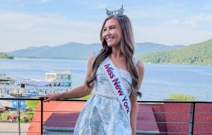 A young woman wearing a tiara, a sash reading Miss New York, a short light blue dress, and white heels stands on a balcony with scenic lake and mountains—Miss New York explores Albany NY by Amtrak.