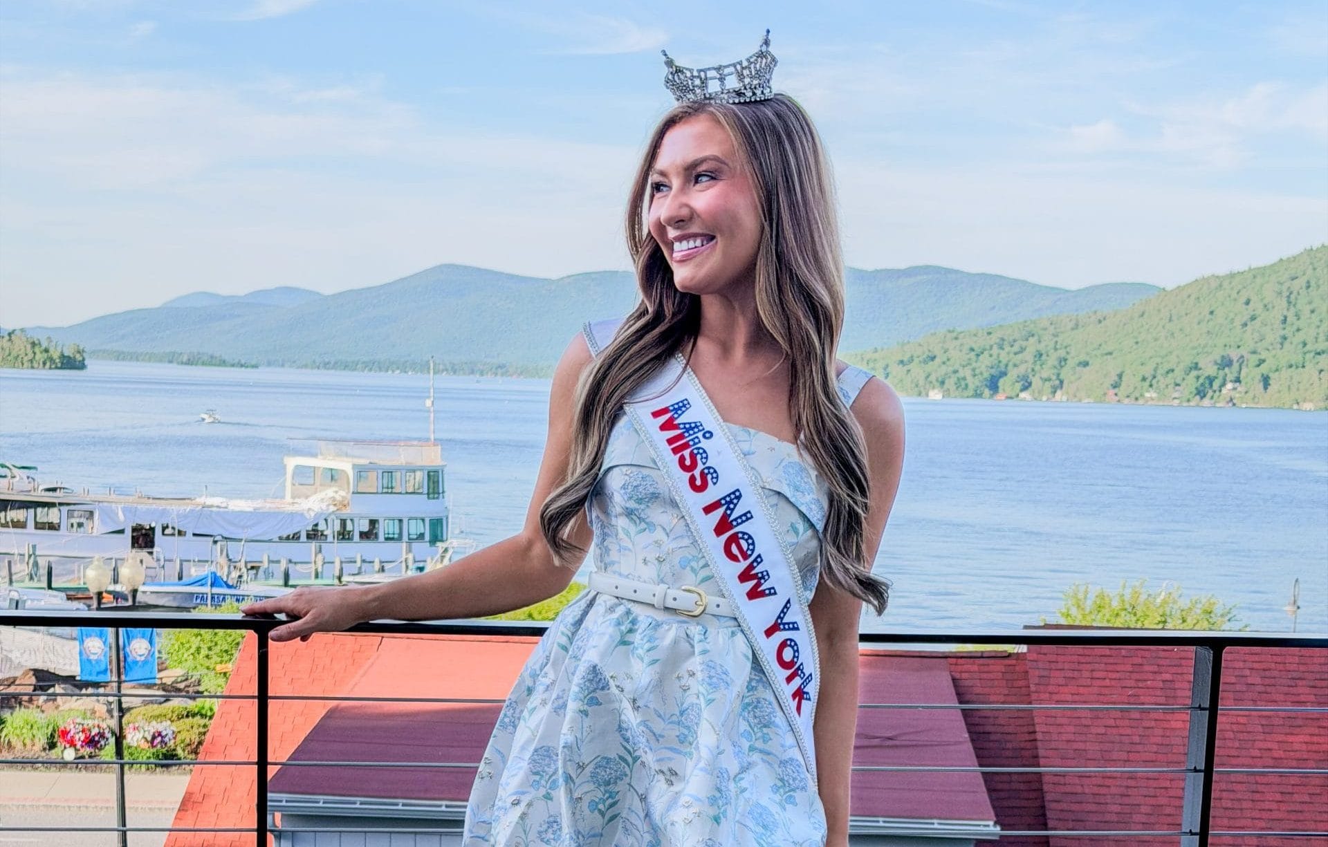 A young woman wearing a tiara, a sash reading Miss New York, a short light blue dress, and white heels stands on a balcony with scenic lake and mountains—Miss New York explores Albany NY by Amtrak.