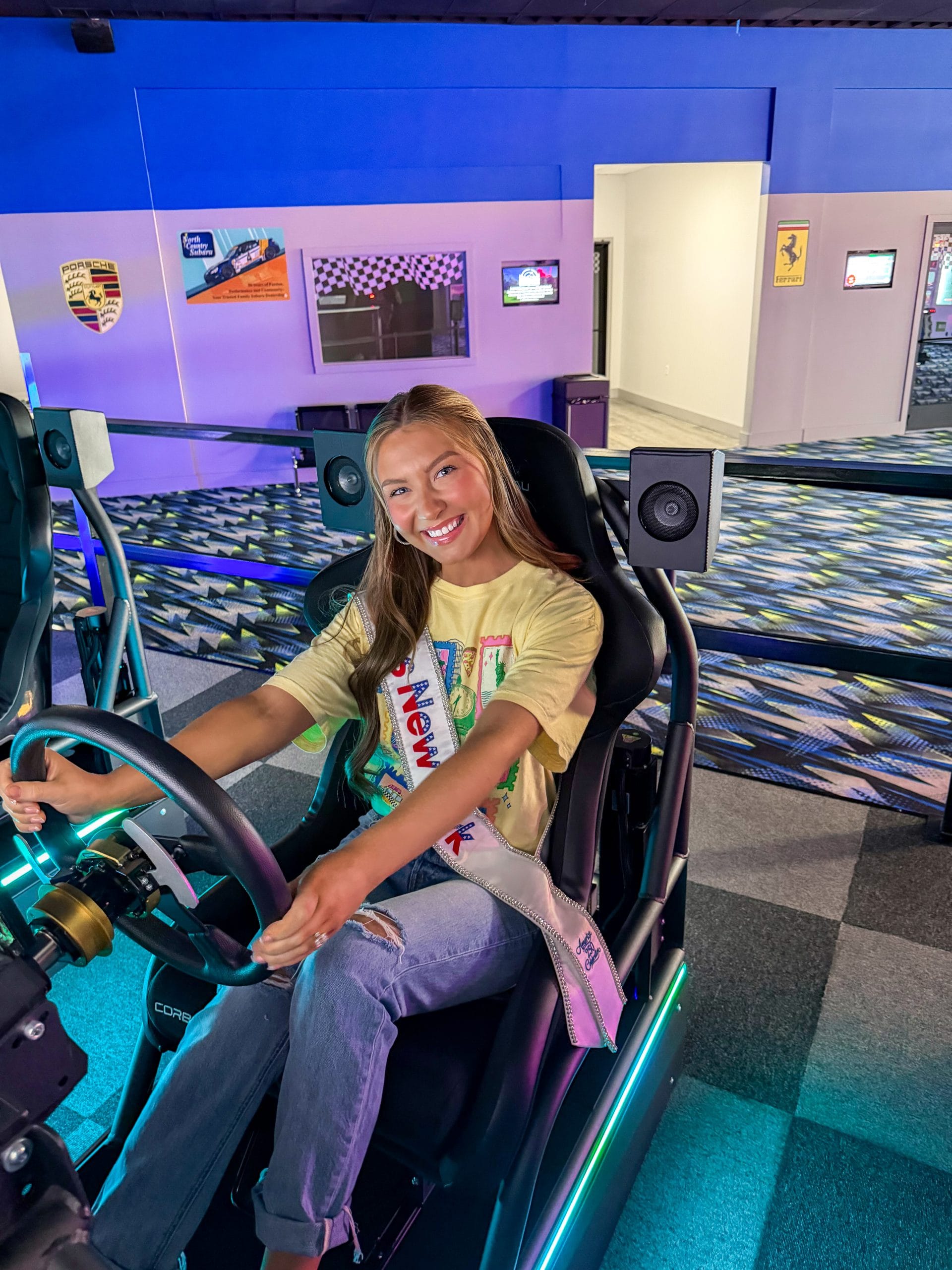 A young woman with long hair, wearing a yellow t-shirt, jeans, and a sash, sits and smiles while holding the steering wheel of a racing simulator in an indoor entertainment center with colorful lighting—an exciting stop near Lake George Area by Rail.