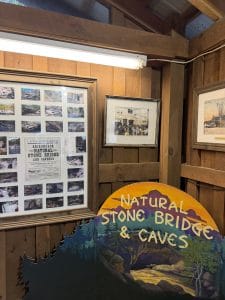 A wooden display wall featuring framed photos and documents, with a colorful sign in front reading Natural Stone Bridge & Caves—decorated with trees and rock formations—highlights the Lake George Area by Rail experience.