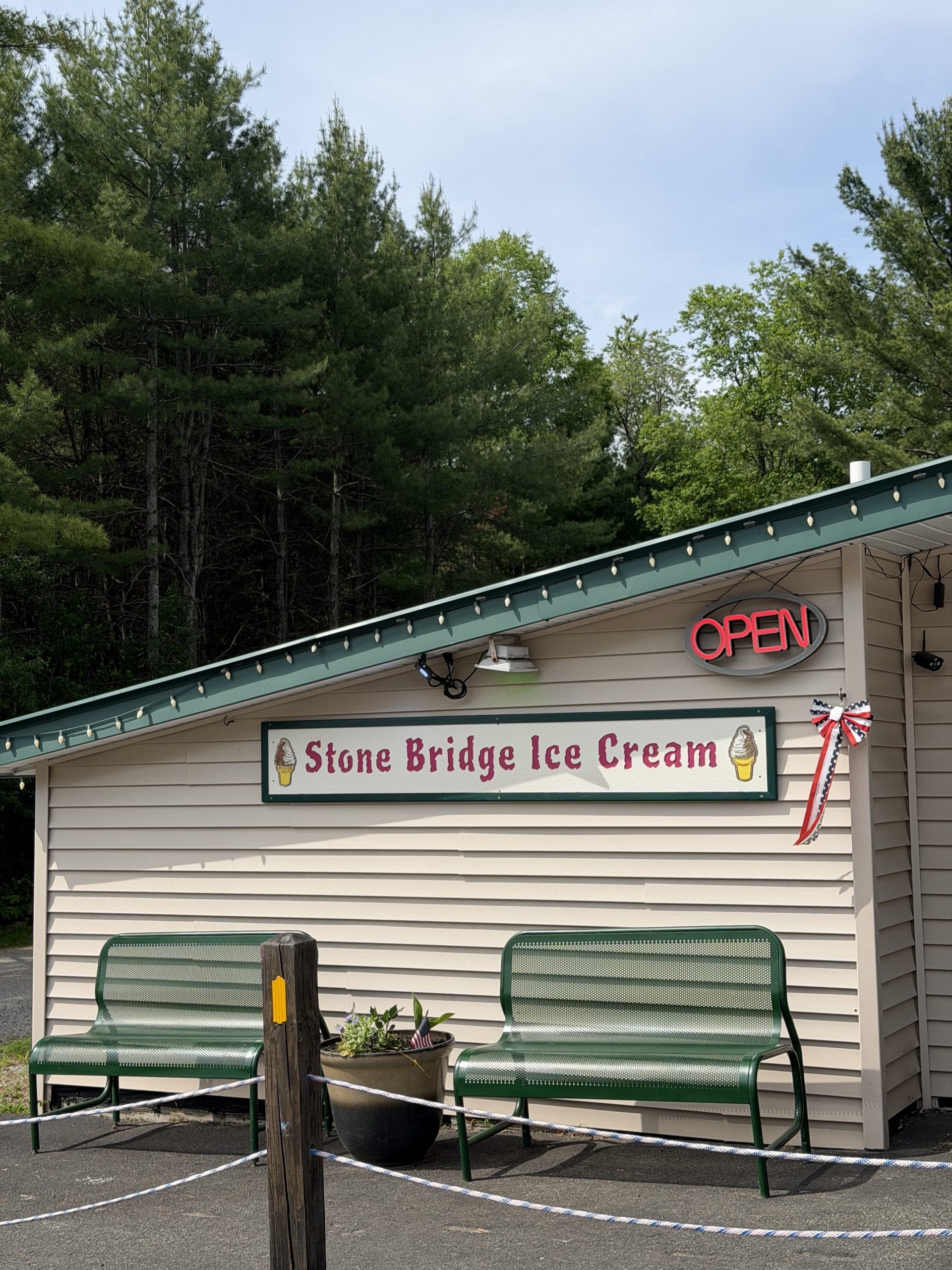 A small beige building with a green roof houses Stone Bridge Ice Cream, a favorite stop for travelers exploring the Lake George Area by Rail. The sign features ice cream cones, and a red neon OPEN glows above two green benches outside among the trees.