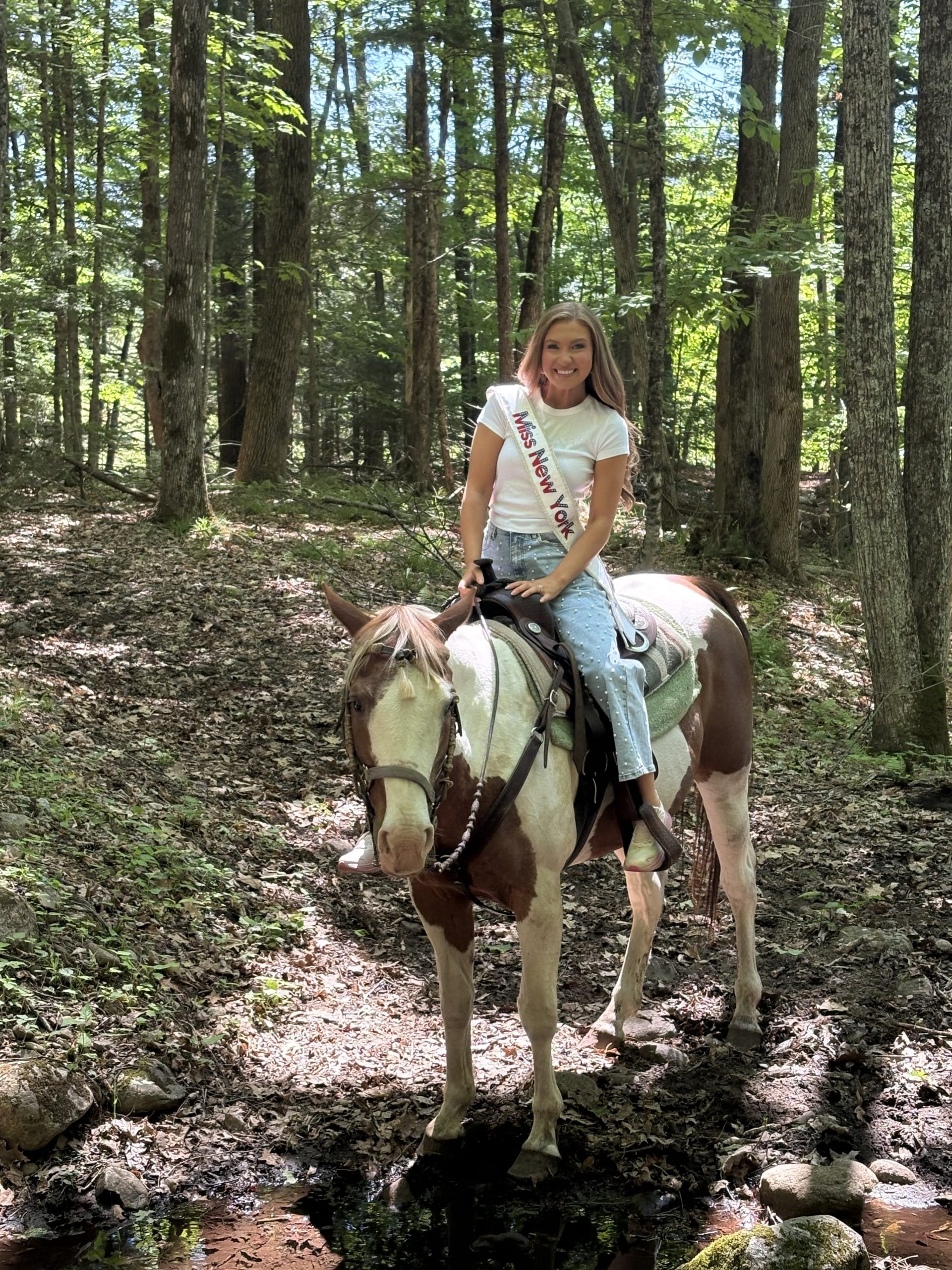 A young woman wearing a sash that reads Miss New York sits on a brown and white horse in a sunlit, wooded forest area near the scenic Lake George Area by Rail, smiling at the camera.