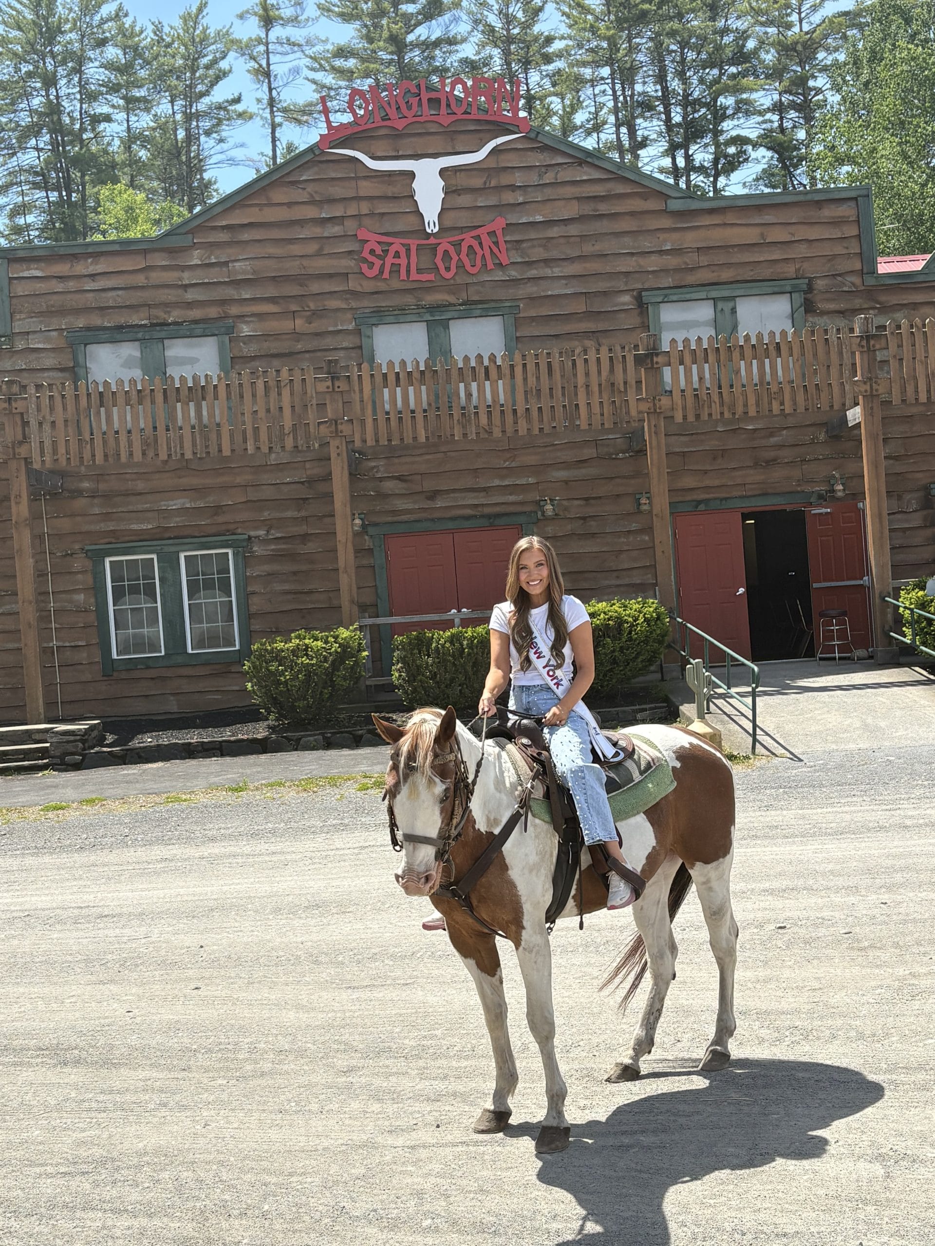 A young girl wearing a sash and jeans sits on a brown and white horse in front of the rustic wooden Longhorn Saloon building, surrounded by trees under a sunny sky—an authentic scene from the Lake George Area by Rail.