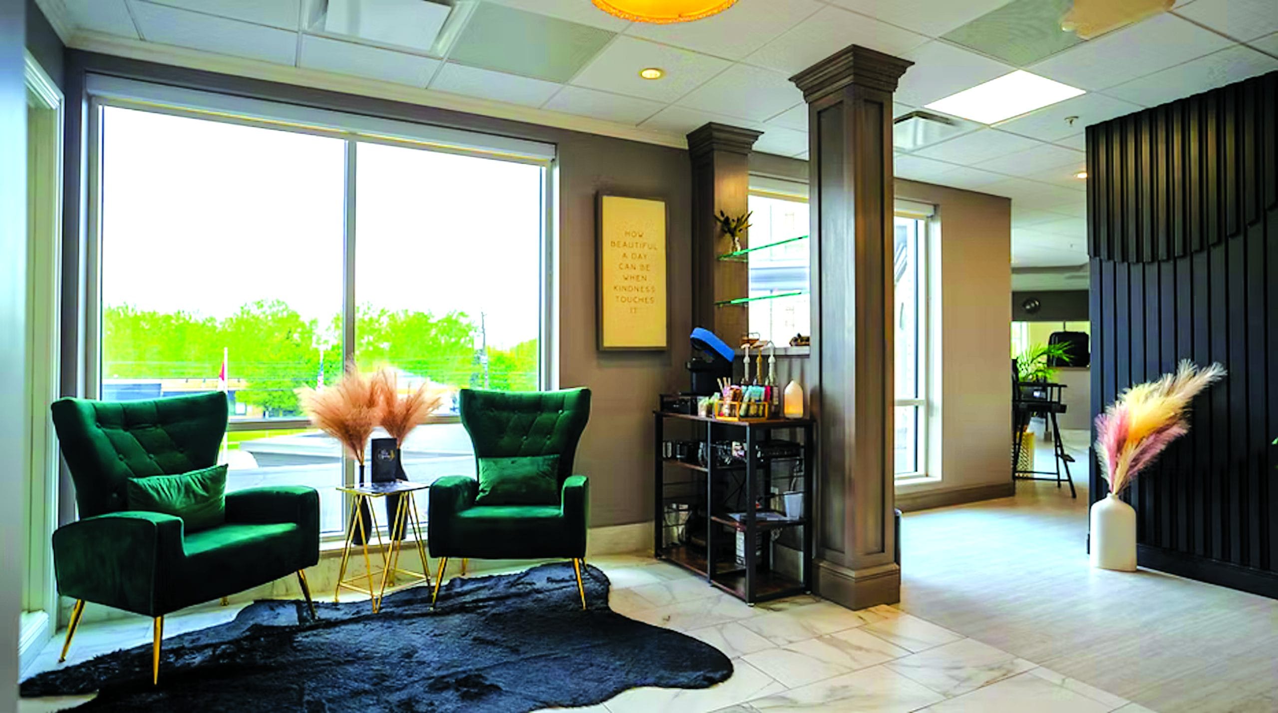 A modern salon lobby with two green velvet chairs, a black rug, a side table with dried pampas grass, and a refreshment cart sets the tone for your Wellness Track. Large windows fill the elegant, neutral-toned space with natural light.