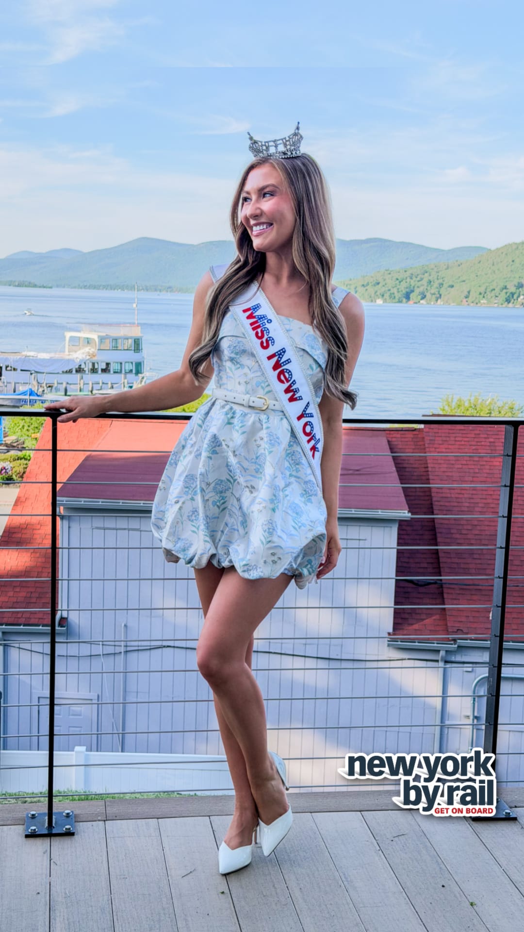 A woman wearing a crown, sash reading Miss New York, and a floral dress stands smiling on a balcony overlooking the Lake George Area by Rail. The newyork by rail logo appears in the corner.