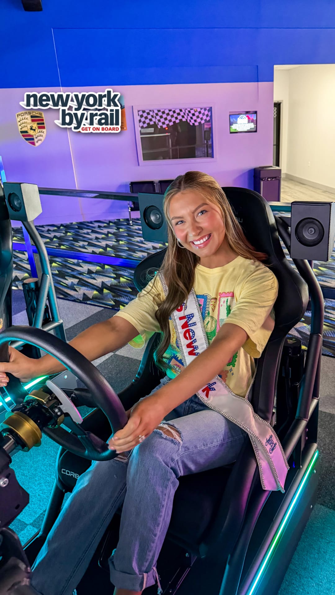 A smiling young woman with long hair sits in a racing simulator, holding the steering wheel. She wears a sash and jeans, with a Lake George Area by Rail sign and checkered decor visible in the background.