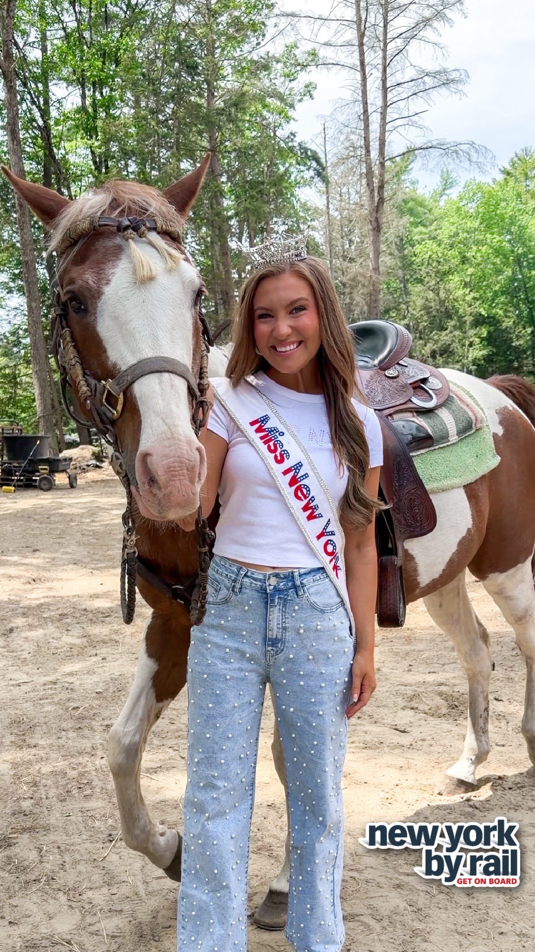 A smiling woman wearing a tiara and a Miss New York sash stands beside a brown and white horse outdoors, dressed in a white t-shirt and pearl-studded jeans. Explore the Lake George Area by Rail! The new york by rail logo appears at the bottom right.