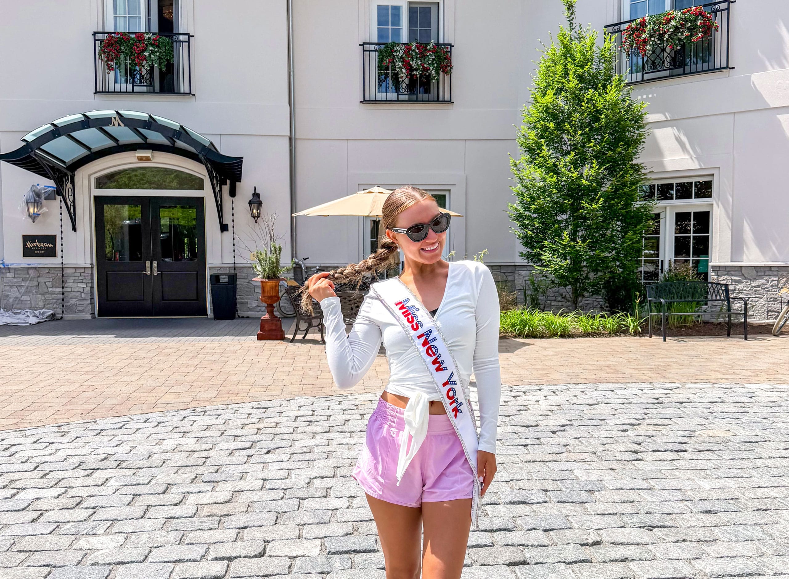 A smiling woman wearing sunglasses, a white sash reading Miss NY, a white top, and pink shorts stands on a cobblestone area in front of the Mirbeau Inn, surrounded by flowers and greenery.