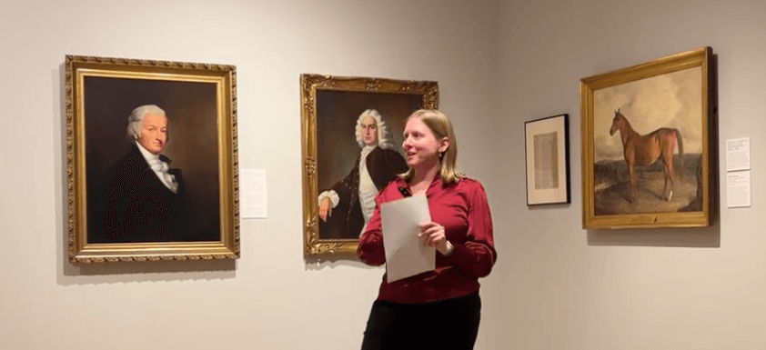 A woman holding papers stands in an art gallery, speaking in front of three framed paintings—including a horse portrait that hints at Racing at the Dawn of the United States.
