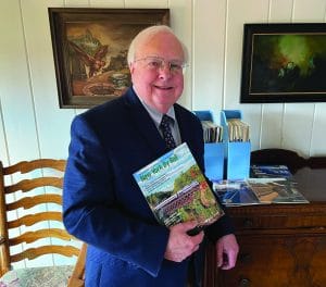 An older man in a suit and tie smiles while holding a “New York By Rail” magazine—perhaps reminiscing about The One That Got Away. He stands indoors beside a wooden table with magazines and file folders, while framed paintings hang on the white wall behind him.