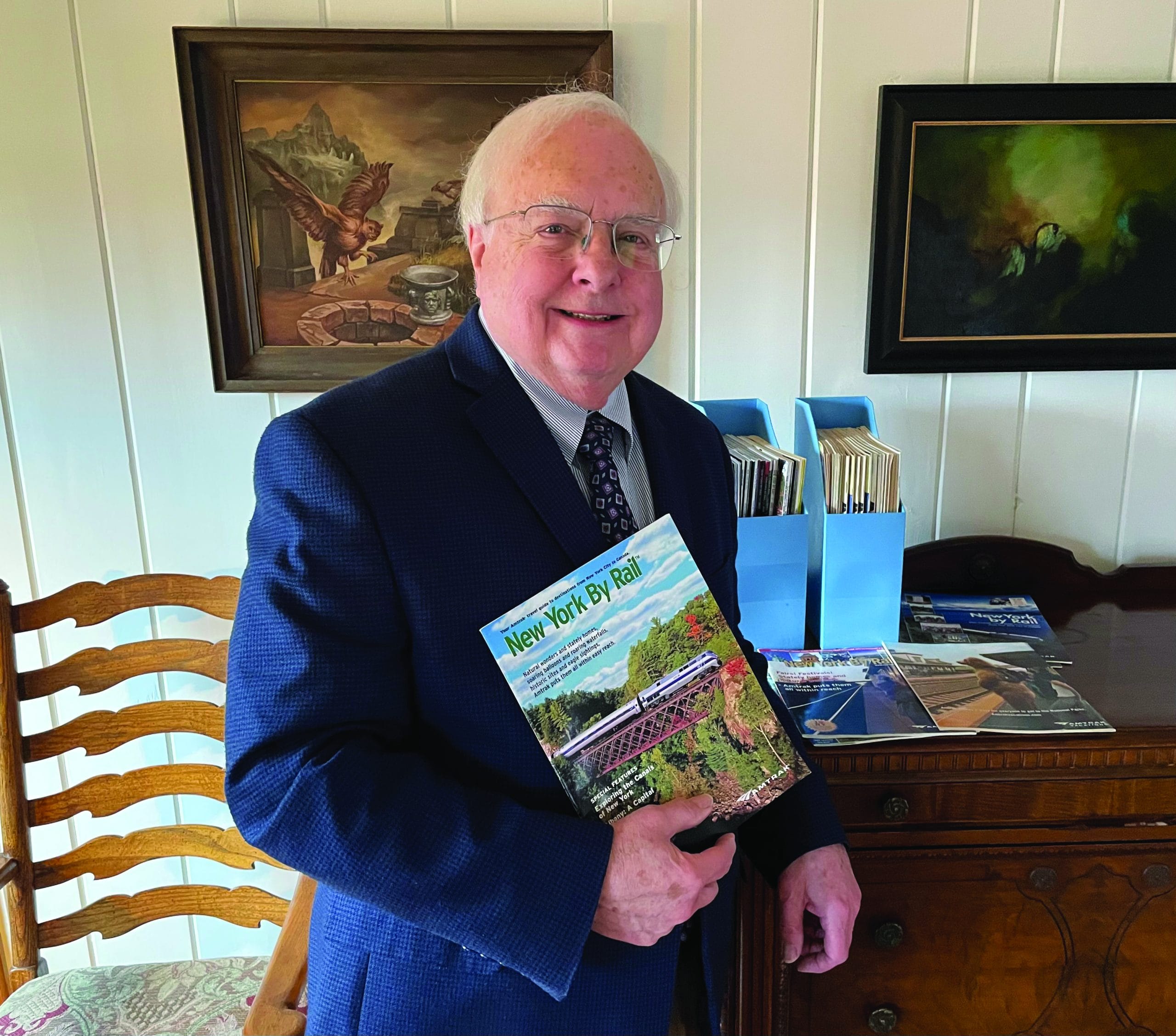 An older man in a suit and tie smiles while holding a “New York By Rail” magazine—perhaps reminiscing about The One That Got Away. He stands indoors beside a wooden table with magazines and file folders, while framed paintings hang on the white wall behind him.