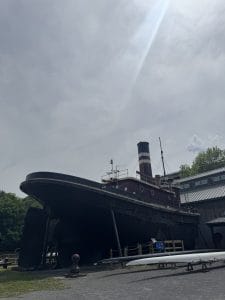 A large, black and red historic tugboat is displayed on dry land outside, with a sunbeam shining from the cloudy sky above and trees and a building in the background—one of the unique free things to do in Albany and Troy.