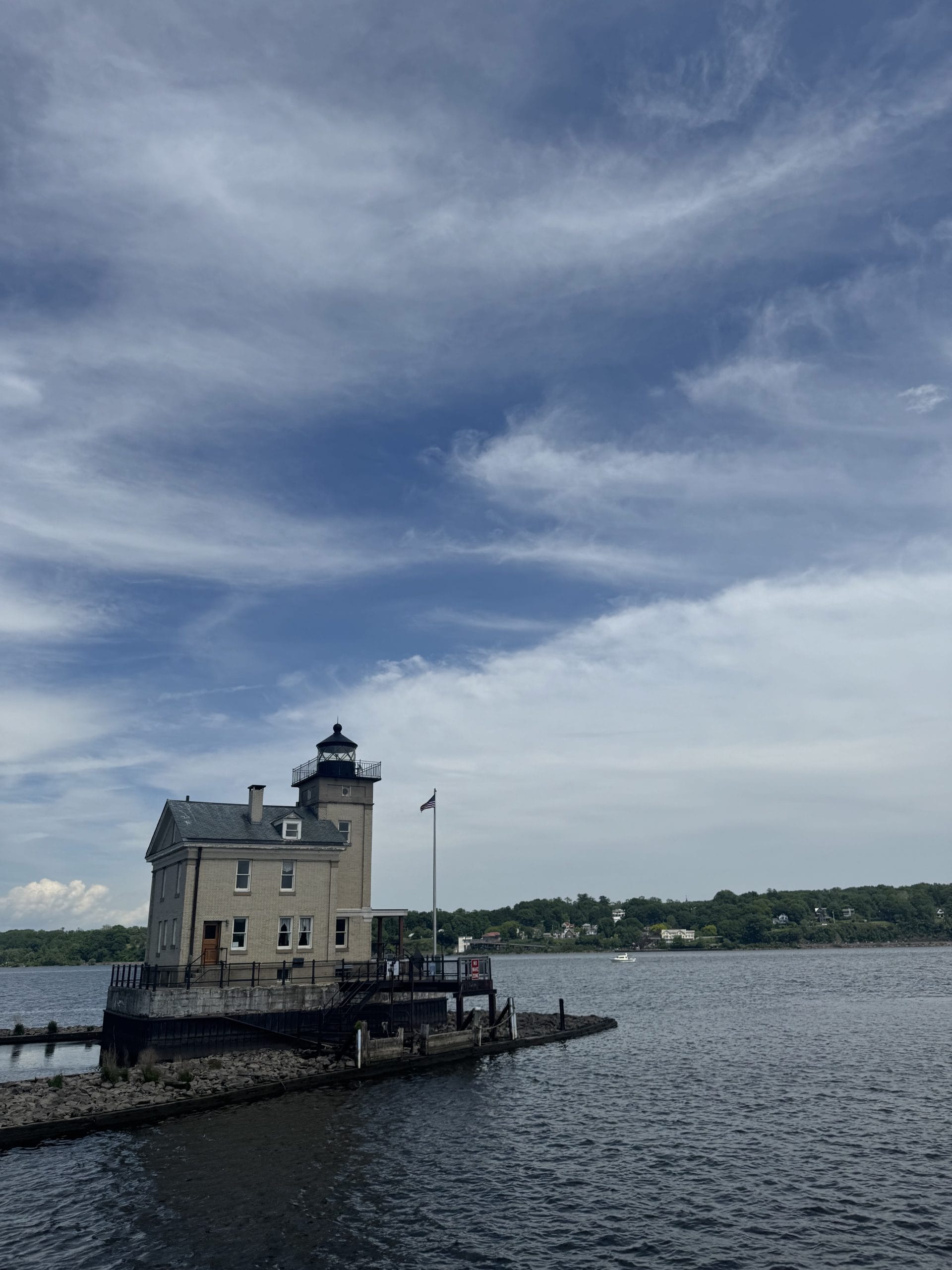 A beige lighthouse with a black lantern stands at the edge of a small pier on a river—one of the free things to do in Albany and Troy—under a partly cloudy blue sky, with green trees and houses on the distant shore.