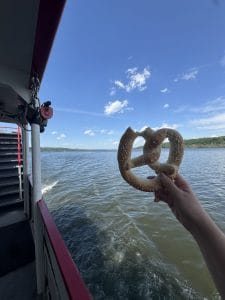 A hand holds a salted pretzel toward the water while standing on a boat, with a blue sky, scattered clouds, and green hills—one of the free things to do in Albany and Troy—visible in the background.