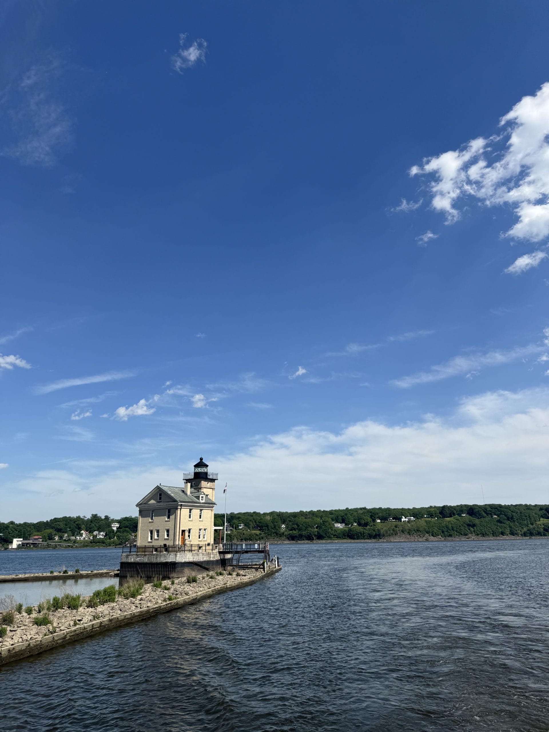 A tan lighthouse with a small house sits at the edge of a stone pier extending into a river—one of the charming free things to do in Albany and Troy—framed by a green tree line and a blue sky with scattered clouds above.