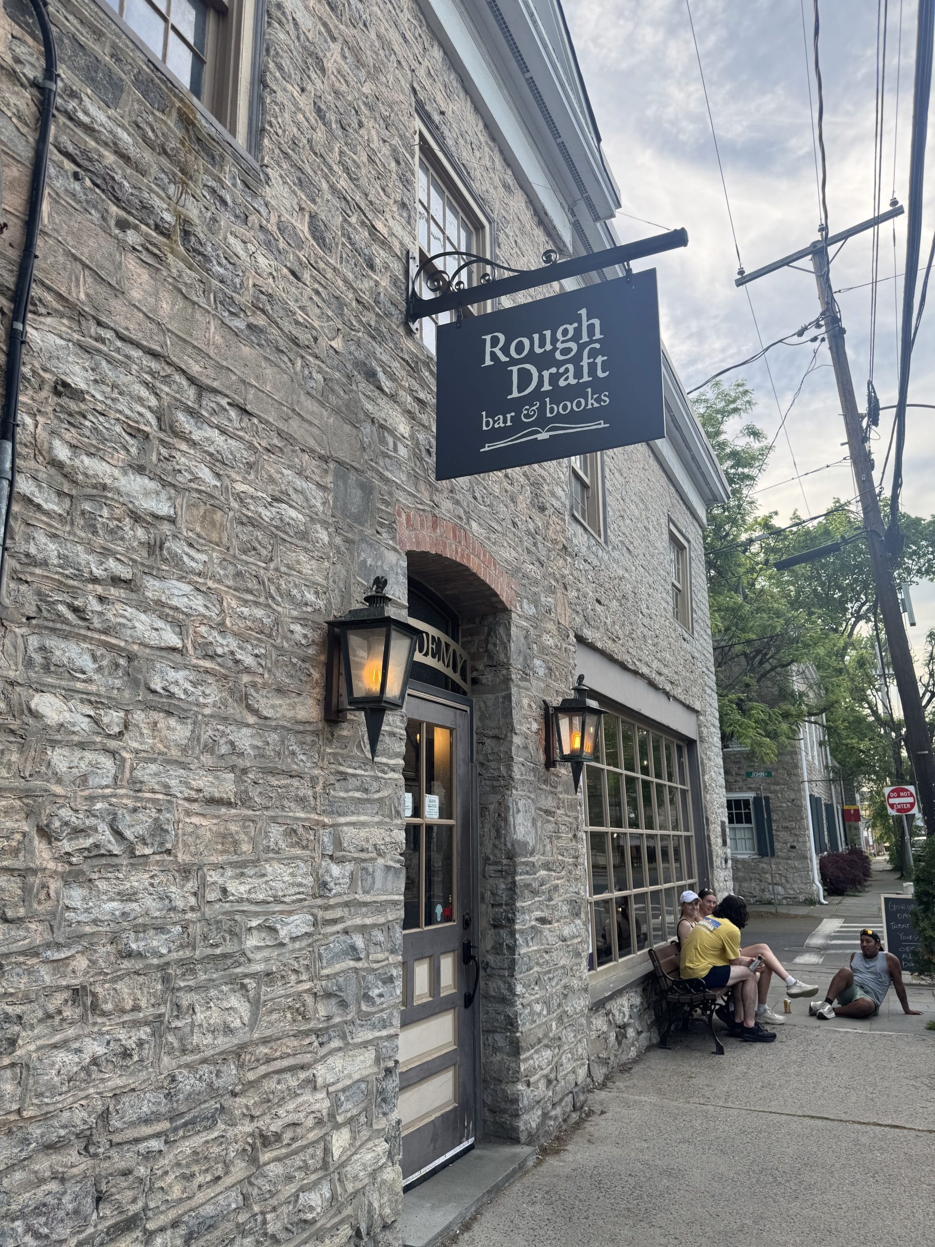 A stone building with a sign reading Rough Draft bar & books hangs above the door. People sit outside on benches, enjoying one of the free things to do in Albany and Troy. Large windows reveal the cozy interior as trees line the sidewalk on a cloudy day.