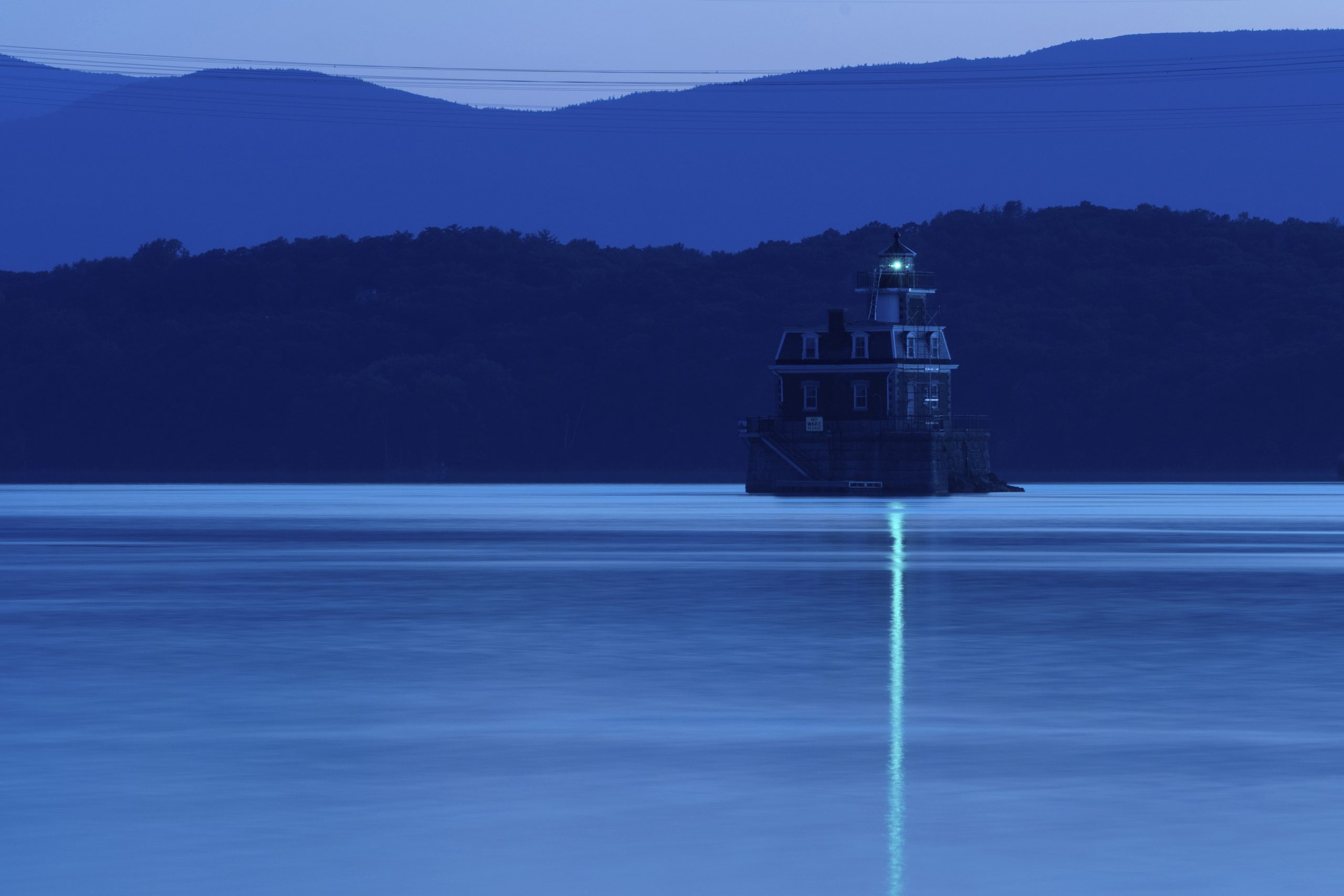A lighthouse with a glowing green light stands on calm water at dusk, surrounded by hills and mountains under a deep blue sky—a tranquil scene reminiscent of the historic Hudson-Athens Lighthouse.